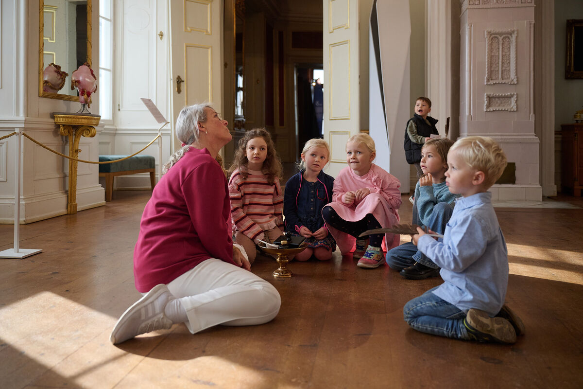Eine Museumsführerin sitzt mit den Kindern auf dem Boden. Sie erzählt, wie Kinder früher im Schloss lebten.